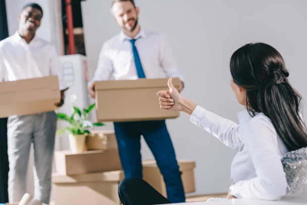 businesswoman showing thumb up to male colleagues holding cardboard boxes in new office - Photo, Image