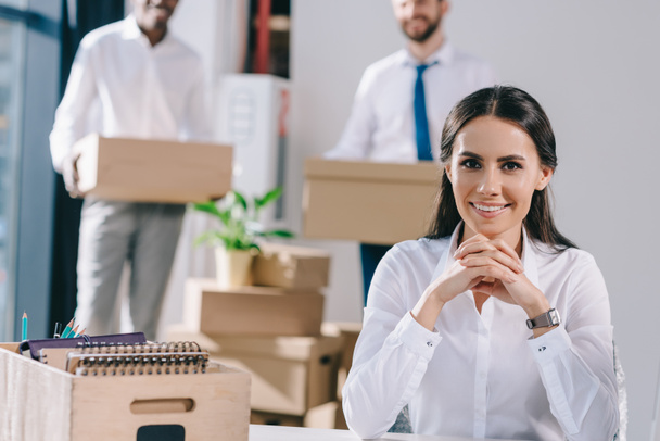 jonge zakenvrouw zitten en glimlachend in de camera terwijl mannelijke collega's houden dozen achter in nieuw kantoor - Foto, afbeelding
