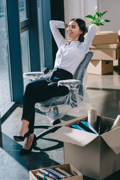  happy young businesswoman sitting with hands behind head in new office - Photo, Image