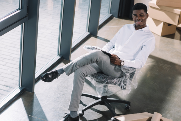 giovane uomo d'affari africano americano con appunti seduto e sorridente alla macchina fotografica nel nuovo ufficio
 - Foto, immagini
