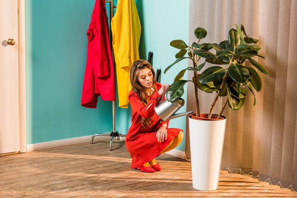 beautiful woman in red dress squatting and watering plant with watering can at home - Photo, Image