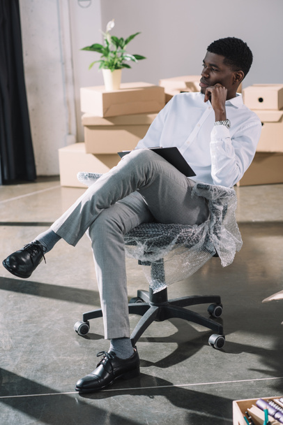 pensive young african american businessman with clipboard sitting and looking away in new office - Foto, immagini