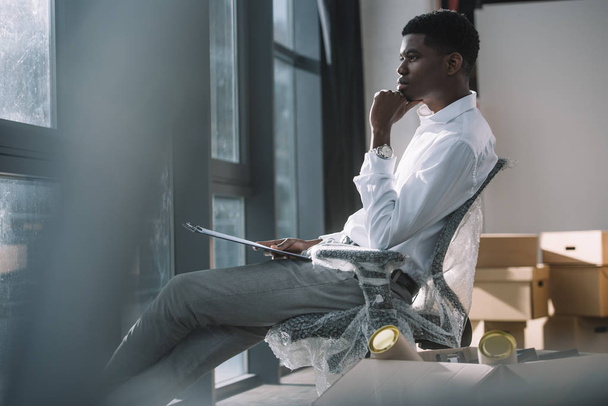 junger afrikanisch-amerikanischer Geschäftsmann hält Klemmbrett und blickt auf Fenster, während er während des Umzugs im neuen Büro sitzt - Foto, Bild
