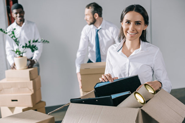 glückliche junge Geschäftsfrau packt Karton mit Büromaterial aus und lächelt in die Kamera, während männliche Kollegen im neuen Büro hinten stehen - Foto, Bild