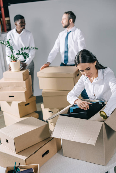 молода бізнес-леді розпаковує картонну коробку і багатоетнічних чоловіків, що стоять позаду в новому офісі
  - Фото, зображення