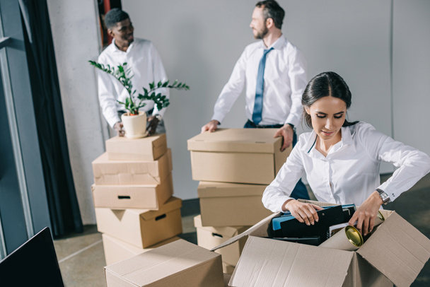 young businesswoman unpacking cardboard box and male colleagues standing behind in new office  - Photo, Image