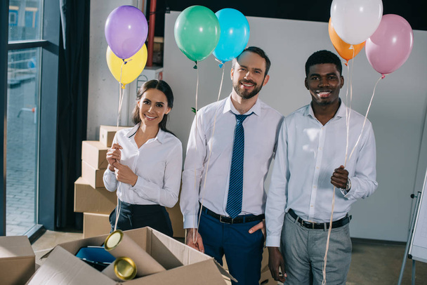 fröhliche multiethnische Kollegen mit bunten Luftballons in der Hand und lächelnd in die Kamera im neuen Büro  - Foto, Bild