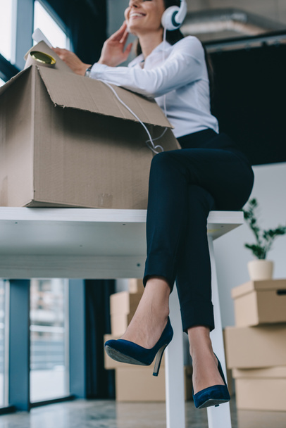 low angle view of smiling young businesswoman in headphones using smartphone while unpacking box in new office   - Photo, Image