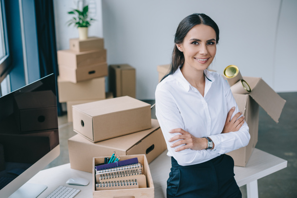 young businesswoman with crossed arms smiling at camera while sitting on table in new office - Photo, Image