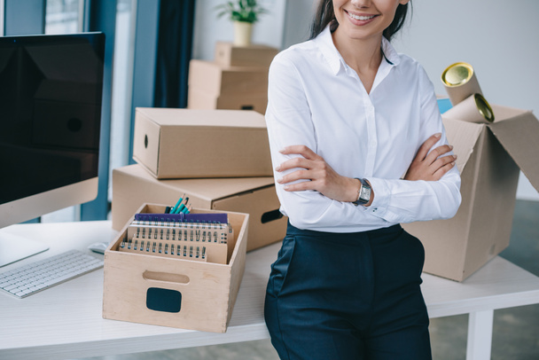 обрезанный снимок улыбающейся деловой женщины со скрещенными руками, сидящей на столе в новом офисе
 - Фото, изображение