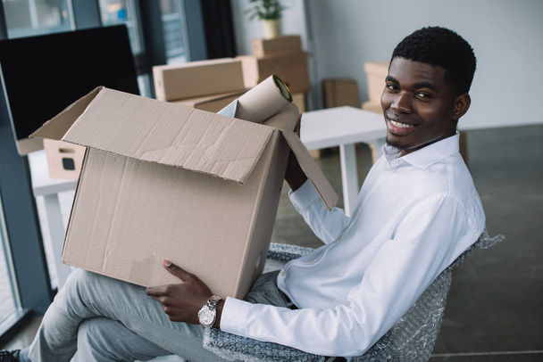 happy african american businessman smiling at camera while holding cardboard box in new office - Photo, Image