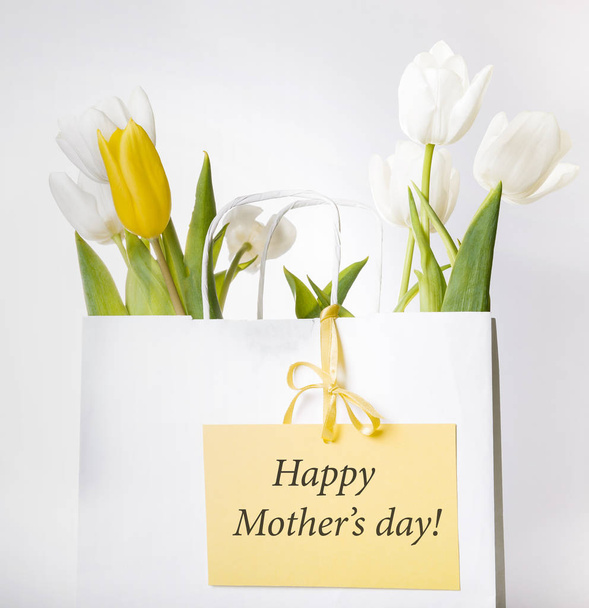 Festive white, yellow tulips composition, handmade heart, ribbon on white background. Bouquet of spring flowers in white paper bag. - Foto, Imagen