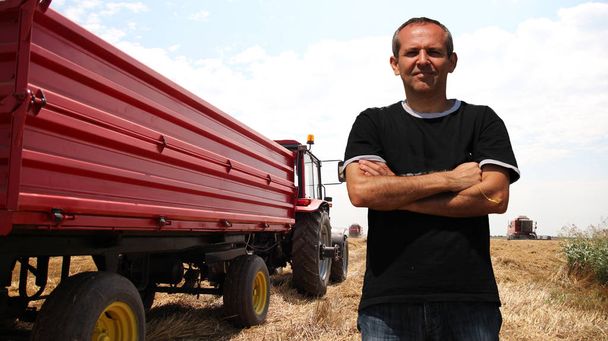 Agricultor en un campo de trigo durante la cosecha / Agricultor de pie frente a un tractor en el campo de trigo
 - Foto, imagen