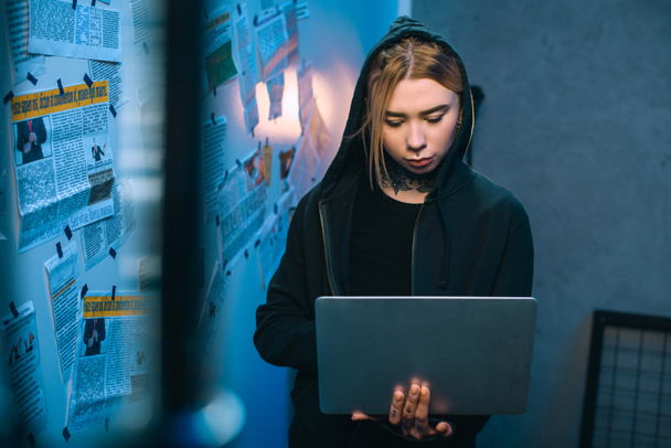 молода жінка хакер з ноутбуком стоячи перед стіну з газетних вирізок - Фото, зображення