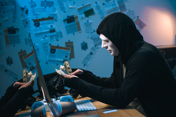 vista lateral de hacker encapuchado en máscara contando dinero robado en su lugar de trabajo
 - Foto, imagen