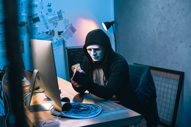 з капюшоном хакер в масці підрахунку вкрадені гроші на своєму робочому місці - Фото, зображення