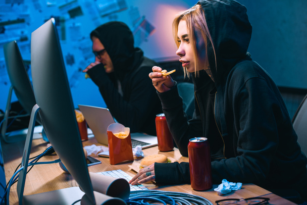 female hacker working on malware with accomplice and eating junk food - Photo, Image