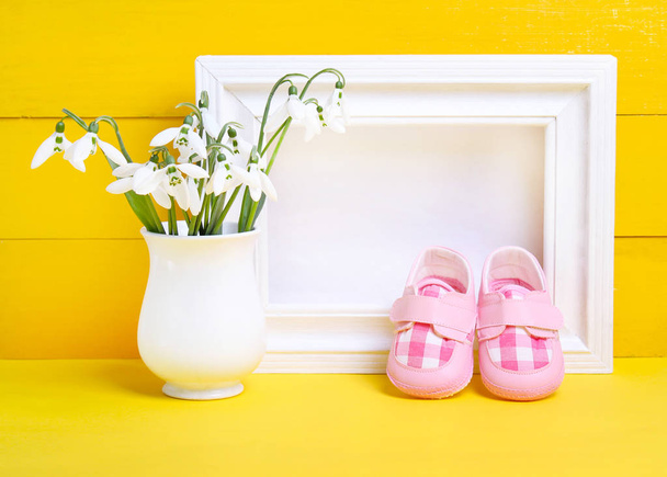 vauvan saappaat, maljakko lumikelloilla ja kehys keltaisella taustalla
 - Valokuva, kuva