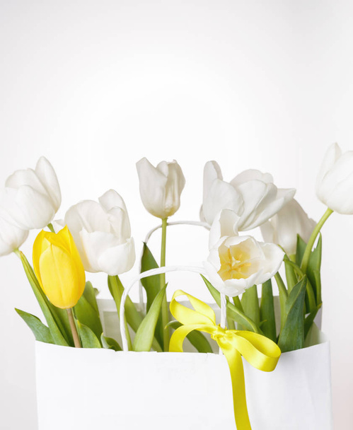 Праздничная белая, желтая композиция тюльпанов, сердце ручной работы, лента на белом фоне. Аромат весенних цветов в белом бумажном пакете
. - Фото, изображение