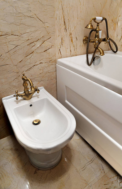 Біде у ванній. Білий біде із золотим батарейним водяним краном та білою ванною всередині ванної кімнати
 - Фото, зображення