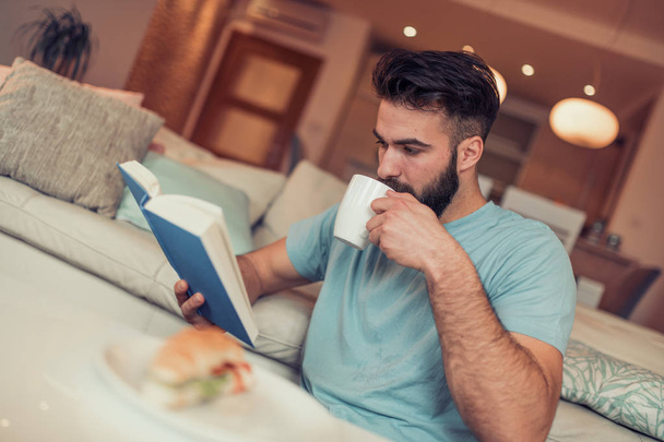 Young man sitting on sofa, reading book and drinking cup of coffee - Foto, afbeelding
