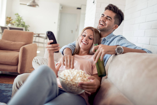 Молодая пара лежит на диване, смотрит кино и ест попкорн с пивом
 - Фото, изображение