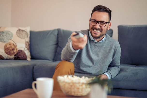 Молодой человек смотрит телевизор с попкорном на диване дома
 - Фото, изображение