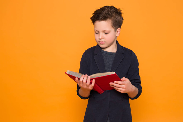 Stay curious! Smart serious little boy wearing suit and reading very interesting book over isolated orange background. - 写真・画像