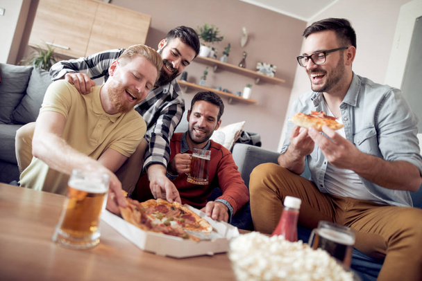 Четверо друзей смотрят телевизор, едят пиццу и пьют пиво дома.
 - Фото, изображение