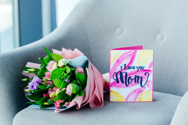 close up view of wrapped bouquet of flowers and i love you mom greeting postcard on armchair, mothers day concept - Photo, Image
