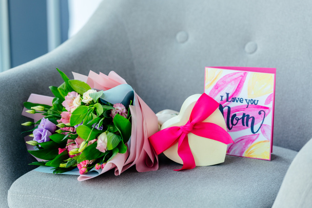 Close-up van verpakte boeket van bloemen, hart gevormde cadeau en ik hou van jou mam briefkaart op leunstoel, moeders dag concept - Foto, afbeelding