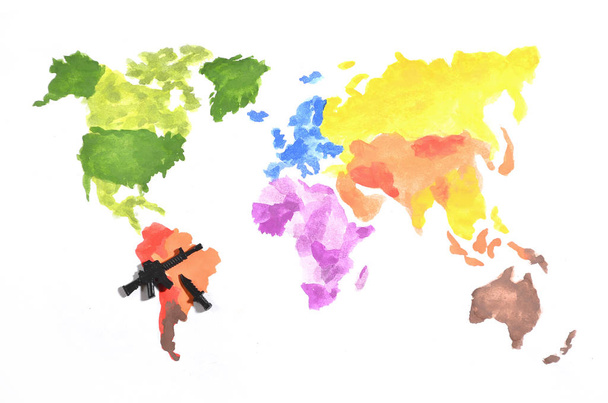 世界地図は、白い紙に黒いおもちゃ銃とナイフの参加で色の水彩絵の具で行われます。南アメリカでの軍事作戦の概念 - 写真・画像
