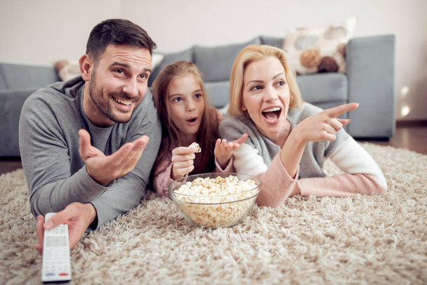 Père, mère et fille couchées par terre, regardant la télé et mangeant du pop-corn
 - Photo, image