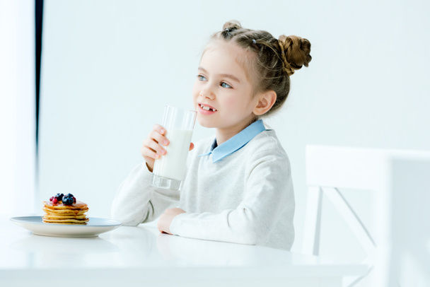 ritratto di bambino carino con bicchiere di latte in mano e frittelle fatte in casa con miele e bacche sul tavolo
 - Foto, immagini