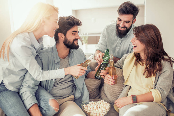 Vrolijke groep vrienden bier drinken en eten popcorn thuis - Foto, afbeelding
