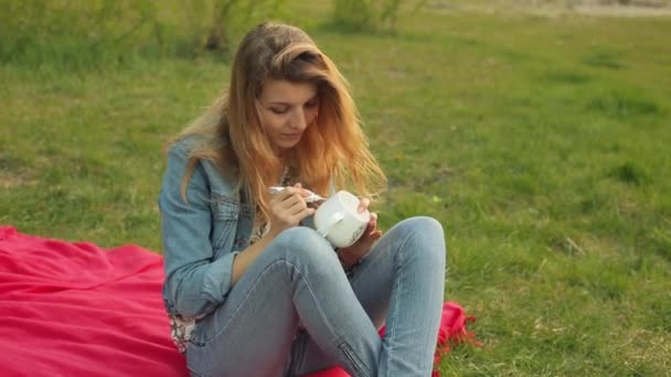 Close-up of female hands drawing a pattern with paint from a tube on a white mug. Girl paints ornament sitting on the red plaid in the evening on the sunset. - Materiał filmowy, wideo
