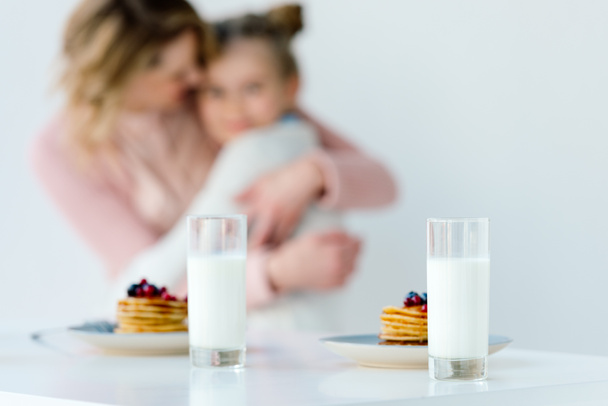 selective focus of glasses of milk and pancakes on tabletop with family hugging behind - Fotó, kép