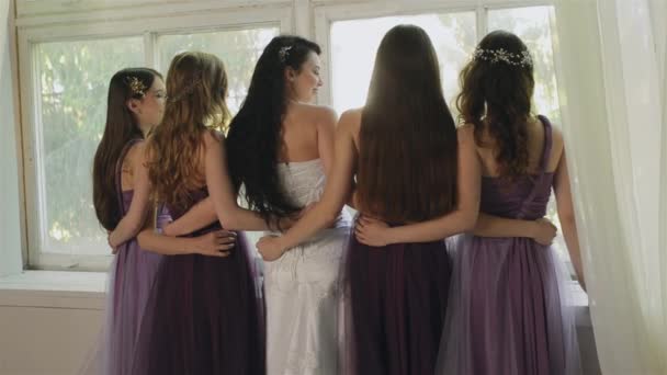 La novia abraza con las damas de honor en el estudio. Hermosas chicas en vestidos púrpura se ríen de la ventana. Mujeres posando durante la mañana de la novia
. - Imágenes, Vídeo