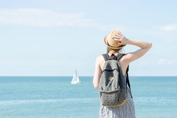 Girl in a hat with a backpack standing on the coastline. Sailboat in the distance. Back view - Фото, изображение