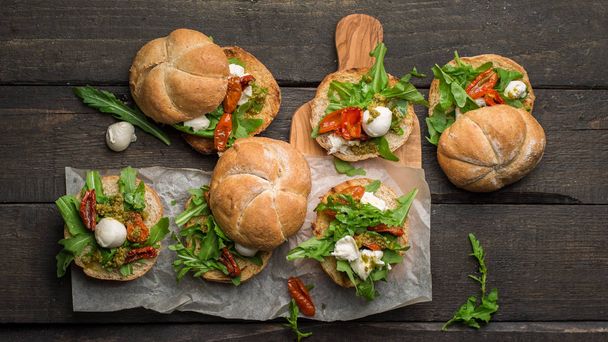 Sandwiches with arugula, dried homemade tomatoes, pesto sauce, mozzarella. Cooking process. Food photo with hands. Atmospheric food photo with wooden dark background. Place for text. Flat lay - Photo, Image