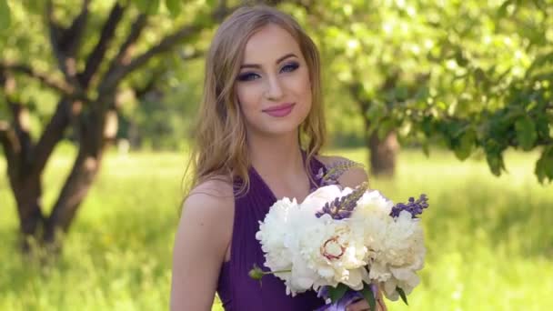 Retrato de una hermosa chica con maquillaje brillante. Mujer rubia caucásica con un ramo de flores posando y mirando a la cámara en vestido lila o púrpura en el jardín
. - Imágenes, Vídeo