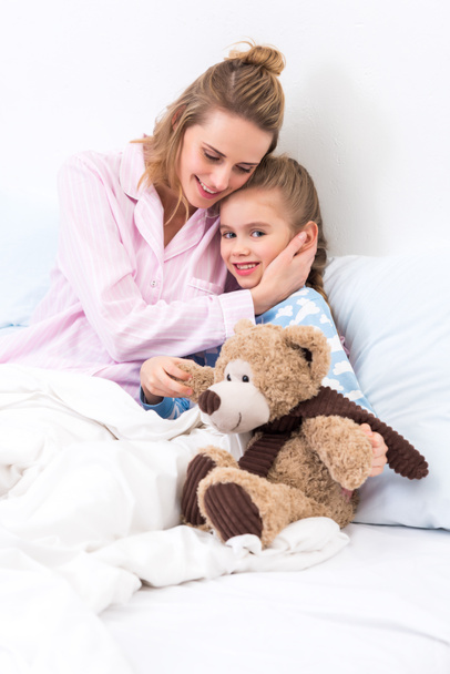 mother hugging daughter with teddy bear on bed at home - Fotó, kép
