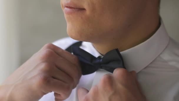 Caucasian man with a short haircut corrects a bow tie near the window in the studio. The groom in a white shirt and butterfly smiles indoors waiting for the bride. - Footage, Video