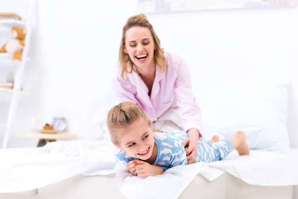 smiling mother tickling daughter on bed at home - Foto, Imagen