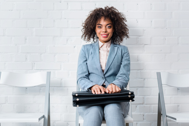 Smiling female candidate with briefcase waiting for interview while sitting on chair - Photo, Image