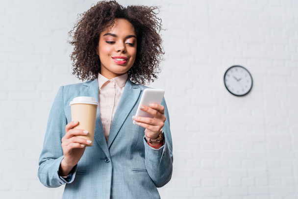 Αφρικανικός Αμερικανός επιχειρηματίας κρατώντας φλιτζάνι καφέ και χρησιμοποιώντας smartphone - Φωτογραφία, εικόνα