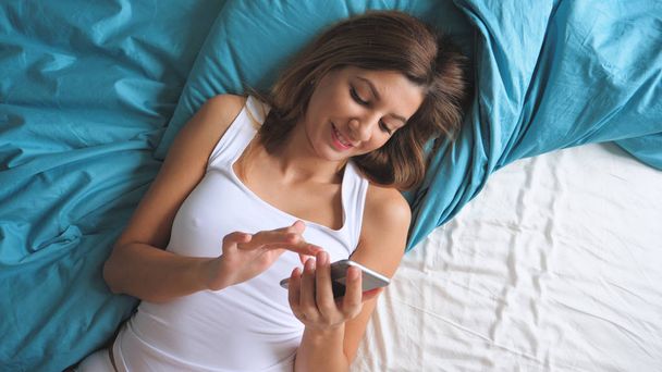 Top View Of Sexy Tanned Woman In Morning Lying On A Bed Uses A Smartphone Apps - Foto, afbeelding