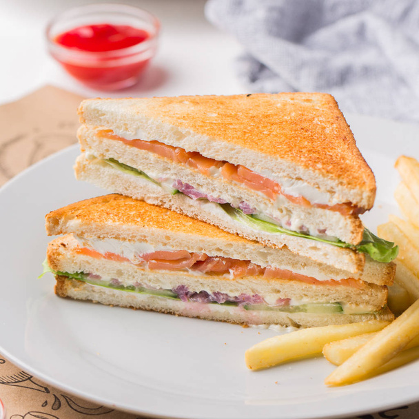 Freshly made club sandwiches with French fries served on white plate.Pressed double toasts with leaf vegetable salad, smoked salmon and cheese served on paper on a wooden table.Street fast food. - Photo, Image