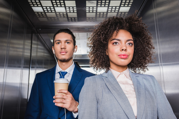 Uomo afroamericano in auricolare con tazza di caffè in piedi in ascensore con giovane ragazza
 - Foto, immagini