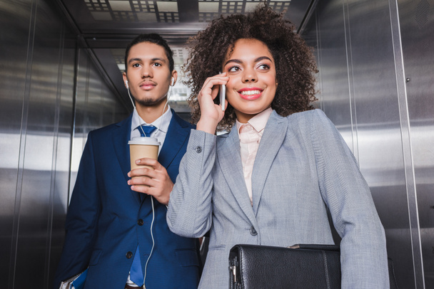 Businessman in earphones with coffee cup standing in elevator with woman talking on phone - Fotó, kép
